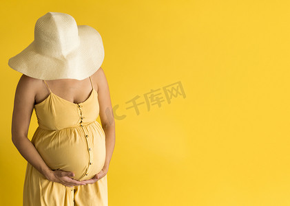 复古摩擦摄影照片_母性、女性气质、时尚、炎热的夏天 — 身穿漂亮复古连衣裙的普罗旺斯草帽、双手放在肚子上、在黄色背景复制空间上摩擦肚子的横幅、无法辨认的孕妇女士