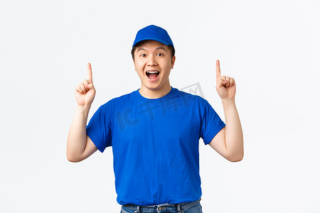 兴奋的微笑亚洲年轻人担任快递员，穿着蓝色制服帽和 T 恤，手指向上，看起来乐观，发布公告，促销报价，白色背景