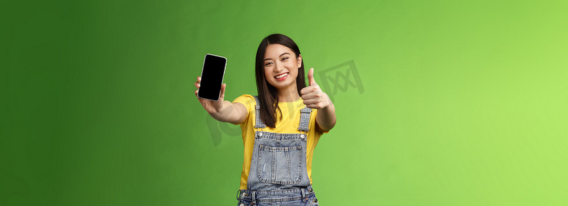 好喜欢摄影照片_欢快满意的可爱亚洲黑发女孩高兴真棒智能手机游戏，显示电话显示屏，竖起大拇指像标志，广泛微笑，介绍真棒在线应用程序，绿色背景