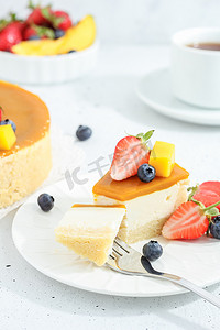 一块芝士蛋糕，盘子里放着芒果，茶叉上装饰着灰色背景上的浆果和鲜花。