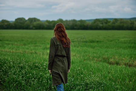 春天多雨的天气里，一位长着美丽红发的女人背对着镜头站在绿色的田野里，穿着长雨衣