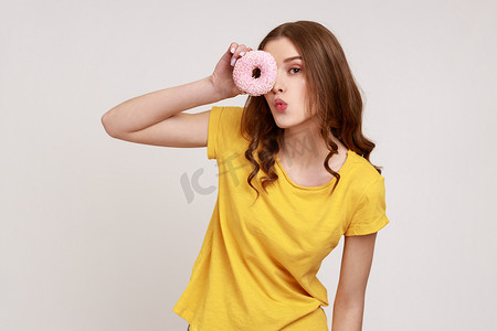 卡通女卷发孩摄影照片_身穿都市风格黄色T恤、积极滑稽的卷发少女的肖像，透过甜甜圈，做出亲吻的手势，玩得很开心。