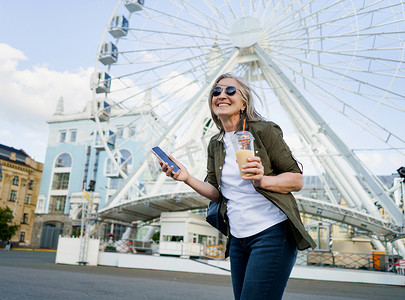 欧洲迷人的灰发女性高兴地享受下班或旅行后的空闲时间，一边拿着手机，一边在城市里使用塑料杯和背景上的摩天轮喝果汁
