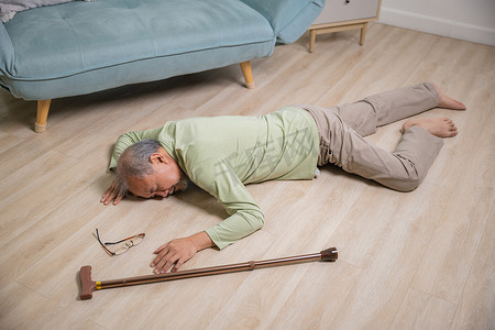 地板摄影照片_亚洲老人独自拿着木拐杖躺在家里的地板上摔倒