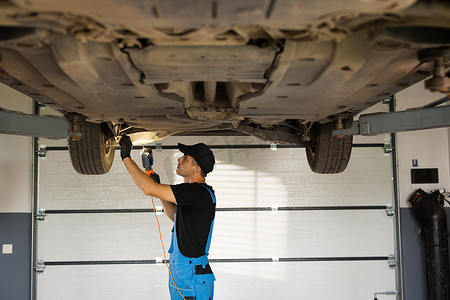 身穿蓝色工作服和黑色帽子的汽车机械师在车库的汽车举升机下工作。