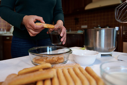 在老式家庭厨房制作传统意大利甜点提拉米苏时，将饼干饼干浸入玻璃杯中，加入浓甜的浓缩咖啡的裁剪图像