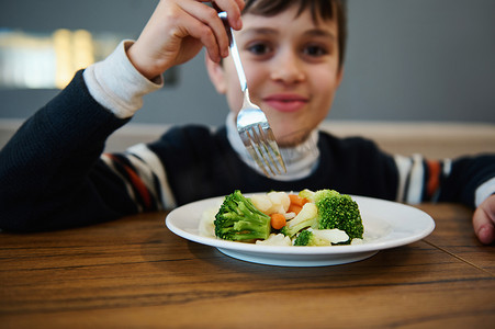 专注于蒸蔬菜、健康的素食食品，在自助餐厅吃午餐时，一个模糊的可爱男孩拿着叉子微笑着看着相机
