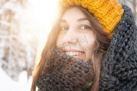 白雪皑皑的森林摄影照片_一位微笑的年轻白人女子的肖像，戴着明亮的羊毛帽子和围巾，背景是白雪皑皑的森林。