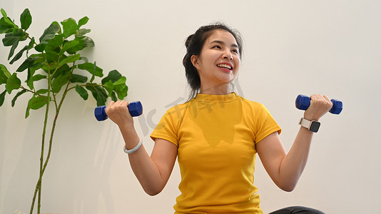 健身教练摄影照片_性格开朗的年轻亚洲女性在明亮的客厅里用哑铃锻炼。