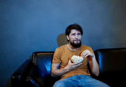 晚上孤独的男人坐在电视机前的沙发上，盘子里放着爆米花