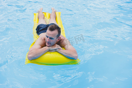一个穿着短裤的年轻人喜欢漂浮在闪闪发光的蓝色水池上的充气大环中的水上公园，对着镜头微笑。