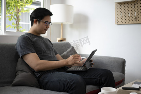 英俊的亚洲男子躺在沙发上，用电脑平板电脑浏览互联网。