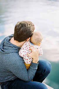 孩子坐爸爸背上摄影照片_爸爸坐在码头上，腿上坐着一个婴儿，看着水面。