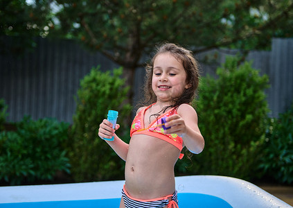 穿着泳衣的可爱小女孩玩得很开心，把肥皂球吹进家里花园的充气池里