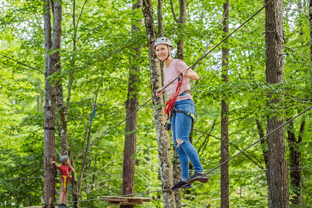 攀岩男孩摄影照片_母亲和儿子在森林里的极限公路电车滑索上攀登树上的登山绳安全连接到树顶绳索冒险公园。