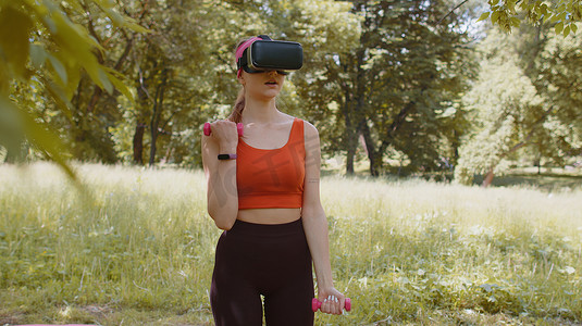 vr头盔摄影照片_戴着 VR 耳机头盔的运动女孩在公园户外用哑铃进行健身锻炼