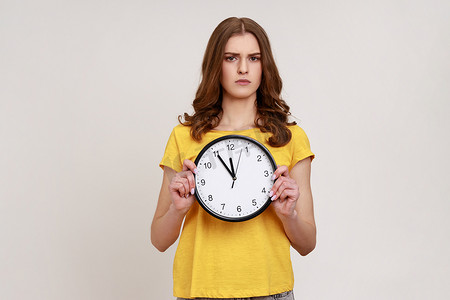 穿着休闲黄色 T 恤、一头波浪发的严肃严格的少女害怕迟到，手里拿着墙上的手表，截止日期，准时。
