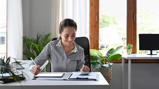微笑的女会计师使用计算器，同时坐在办公桌前，拿着数字平板电脑和财务文件。