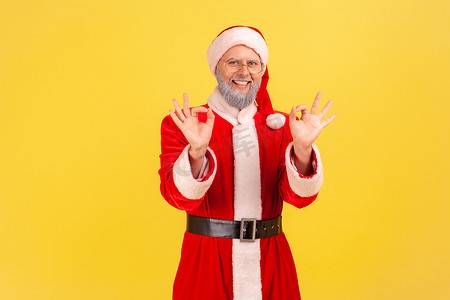 满意的老人的肖像，留着灰胡子，穿着圣诞老人服装，双手显示出好的迹象，看着相机露齿微笑。