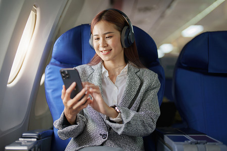 手机播放摄影照片_一位成功的亚洲女商人在飞机上的肖像坐在商务舱座位上，在飞行过程中使用智能手机播放音乐，relex 概念