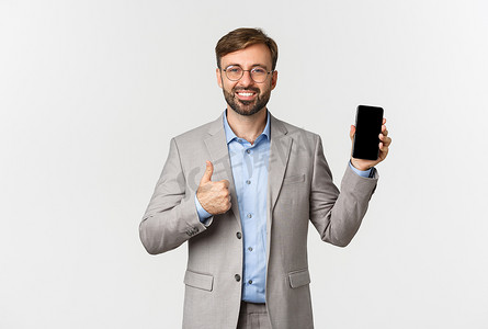 英俊的微笑商人，留着胡子，穿着灰色西装和眼镜，竖起大拇指，在手机上展示一些东西，推荐应用程序，白色背景