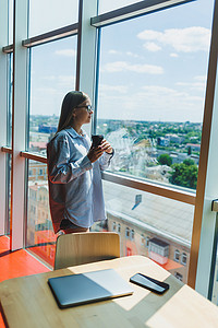 这扇摄影照片_一位戴着眼镜的年轻女性自由职业者拿着咖啡站在一扇大窗户旁，看着这座城市。
