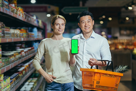 超市里年轻家庭的不同购物者微笑着看着相机、杂货部、男人和女人拿着购物篮，展示智能手机的绿屏