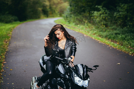 一位美丽的年轻女子骑着摩托车，穿着黑色皮衣，在大自然、街道、森林里，在阳光明媚的天气里，特写，看着相机的肖像