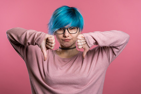 年轻漂亮的蓝头发女人站在粉红色的工作室背景上表达不满，并对着镜头表现出拇指朝下的手势。