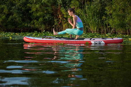 年轻女子正在美丽的湖泊或河流上的立式桨板 SUP 上做瑜伽
