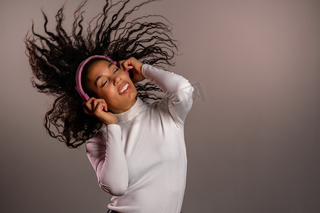 粉红kt板摄影照片_漂亮的混血女孩，一头非洲卷发，在灰色背景的工作室里玩得很开心，微笑着，戴着耳机跳舞。
