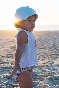 戴着巴拿马帽的金发小女孩站在海滩上，看着大海