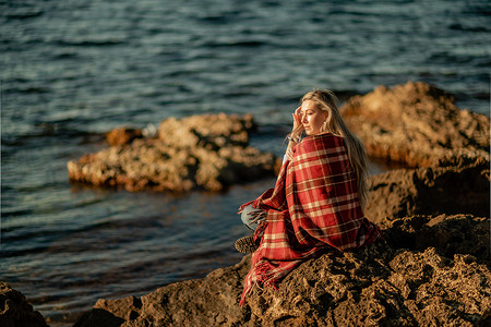 迷人的金发白种女人在日落时在海滩上享受时光，坐在毯子里，看着旁边，背景是日落的天空和大海。