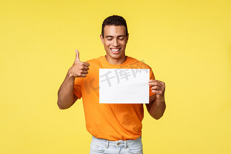 穿着橙色 T 恤的快乐西班牙裔帅哥，拿着白纸，看着那张纸，微笑着表示赞同，表现出竖起大拇指的手势，比如促销，建议访问公司网页，黄色背景