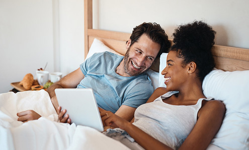 电脑电影摄影照片_周末在家里的室内，笑着的已婚夫妇带着平板电脑躺在床上一起观看有趣的喜剧电影或电影。