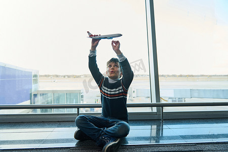 可爱的小男孩坐在全景窗户的背景下玩玩具飞机，俯瞰机场出发航站楼的跑道