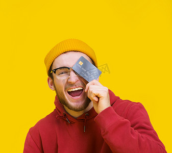 花钱买快乐摄影照片_手里拿着信用卡或借记卡的年轻人高兴地微笑着遮住眼睛准备花钱。