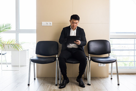 亚洲商人在候诊室坐在办公中心接待处附近的椅子上，填写测试表以接收申请