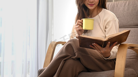 微笑的年轻女子喝着花草茶，坐在扶手椅上看书，在家度过休闲周末