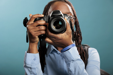 单反图片摄影照片_工作室拍摄的年轻成年女性用单反相机站在蓝色背景上拍摄照片。