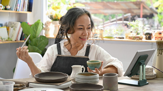 快乐的退休妇女在她的工作室里画陶碗并使用数字平板电脑。