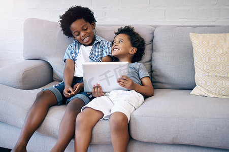 两个可爱的非洲裔美国小男孩舒适地坐在沙发上，使用无线平板电脑，一边浏览和微笑。
