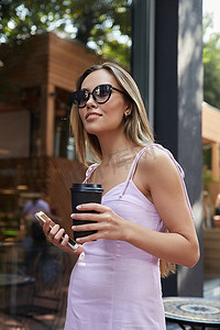 身穿粉色连衣裙的亚洲女性站在户外，手里拿着智能手机和咖啡