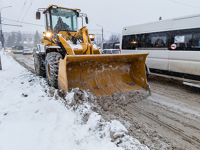 2020灯光摄影照片_俄罗斯图拉 — 2020年11月21日：冬日灯光下，拖拉机用大铲子清理道路上的积雪。