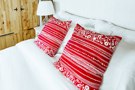 红色毛毯和枕头摄影照片_卧室里的红色枕头配有白色床单和灯