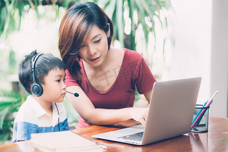 年轻的亚洲母子在家里使用笔记本电脑一起学习，男孩戴着耳机进行电子学习，保持距离，老师或妈妈支持孩子，教育理念。