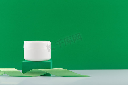 绿色讲台上的白色奶油、面膜或香膏罐，绿色背景上有丝带，有复制空间
