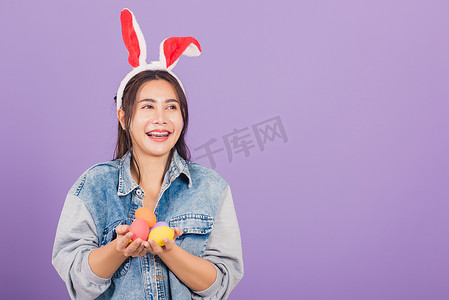 耳钉摄影照片_戴着兔耳和牛仔裤微笑的女人手上拿着彩色复活节彩蛋礼物