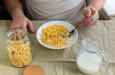 一个剪短的男人拿着一把勺子，里面装着玉米片，里面装着牛奶，盘子里装着牛奶，盘子放在朴素的粗糙桌布上。