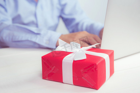 男人的手拿着红色礼品盒，木桌上放着笔记本电脑，网上购物为圣诞节或周年纪念日送礼物，在社交距离期间庆祝和庆祝。